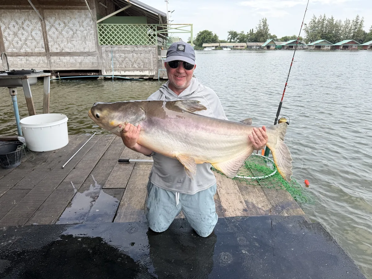 An angler kneeling on a mat and holding a pale catfish beside the lake