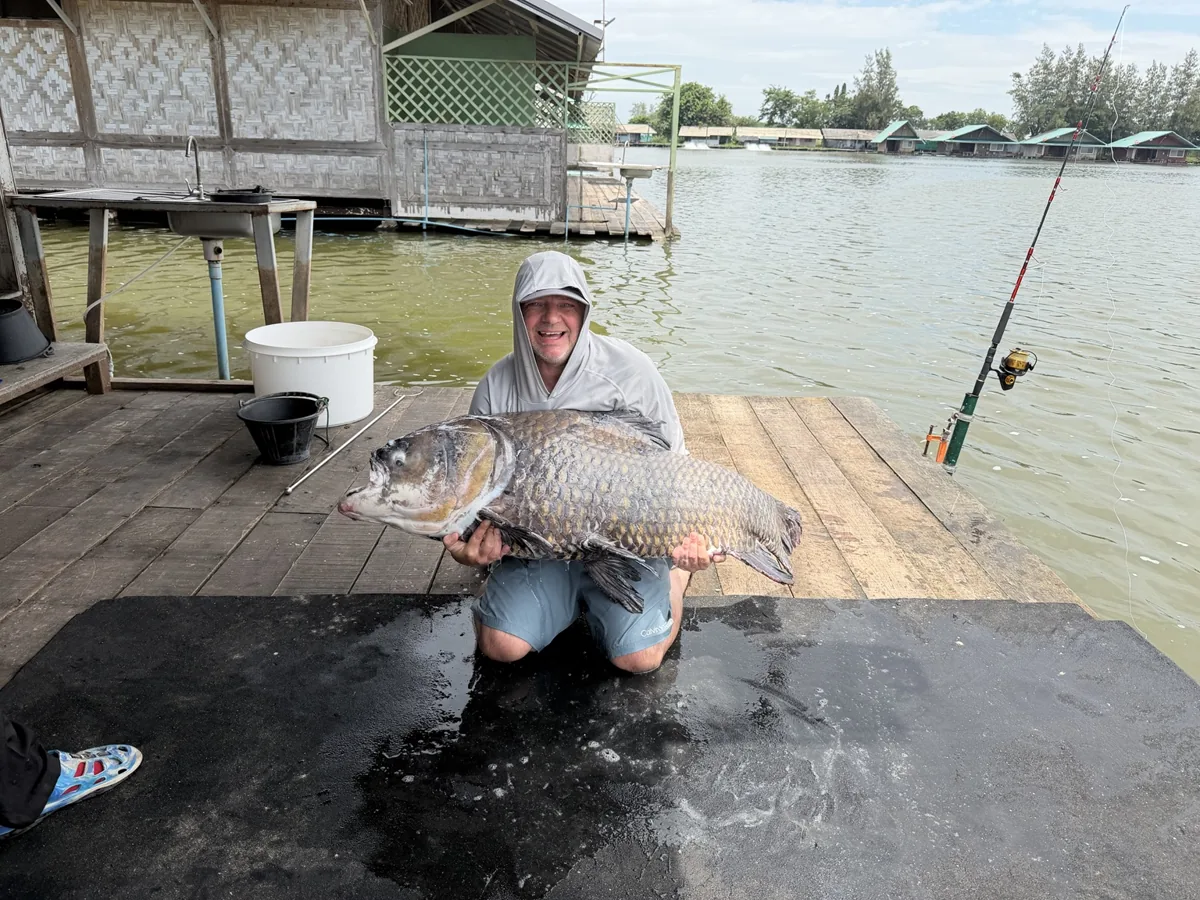 An angler kneeling on a black mat with a large scaled fish beside a lakeside structure