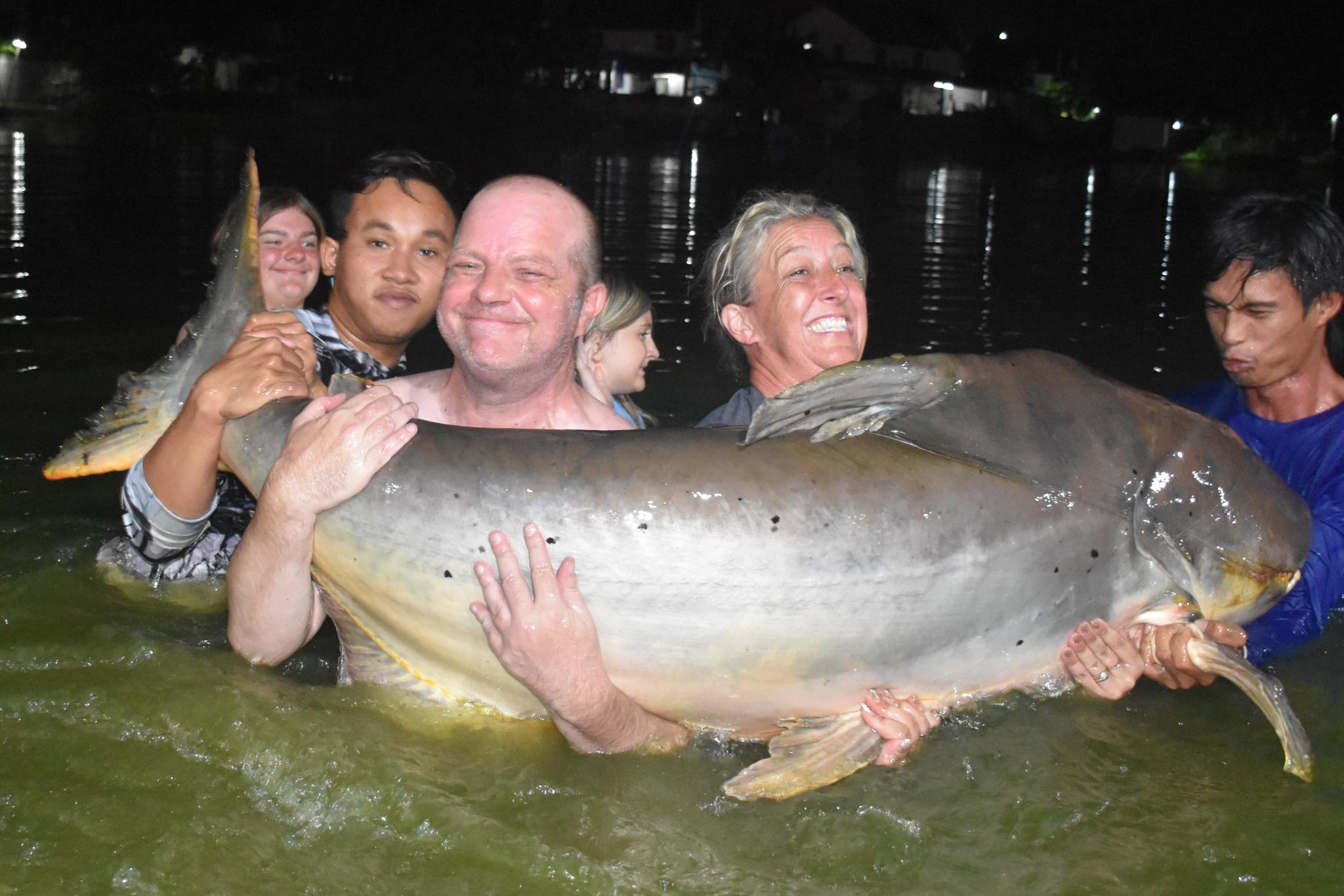 A group supporting a very large catfish in the water at night