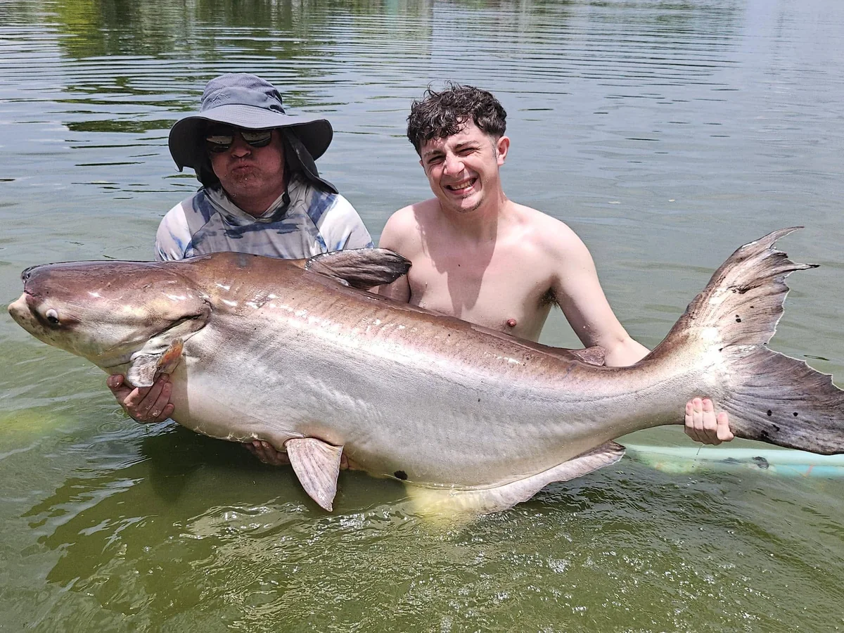 Finlay and a helper supporting a large catfish in the lake.