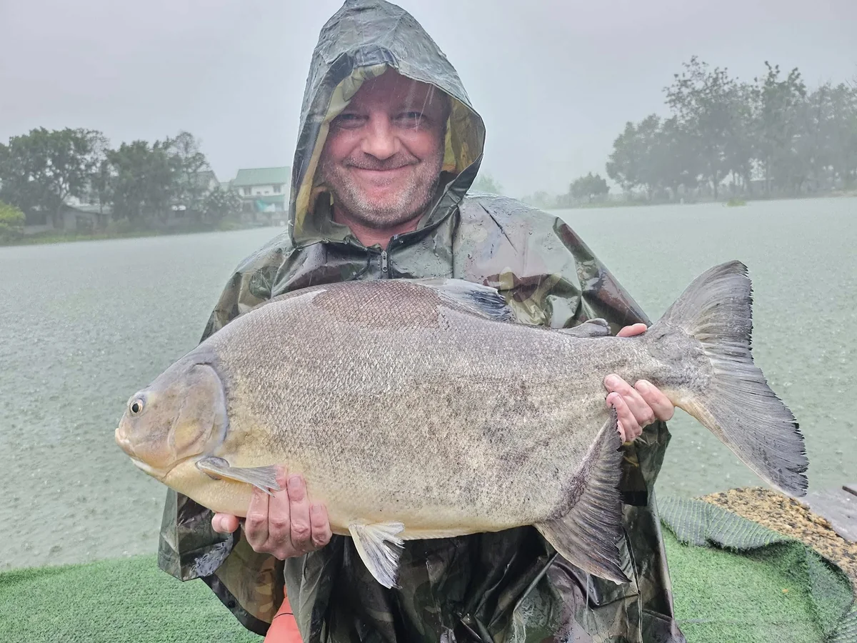 Vinny in a hooded waterproof holding a broad-bodied fish beside the lake in heavy rain.