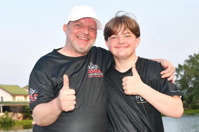Vinny and Tyler giving thumbs-up in matching catfish club shirts beside the lake.