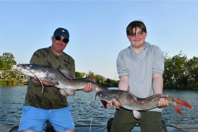 Vinny and Tyler each holding a catfish together beside Palm Tree Lagoon.