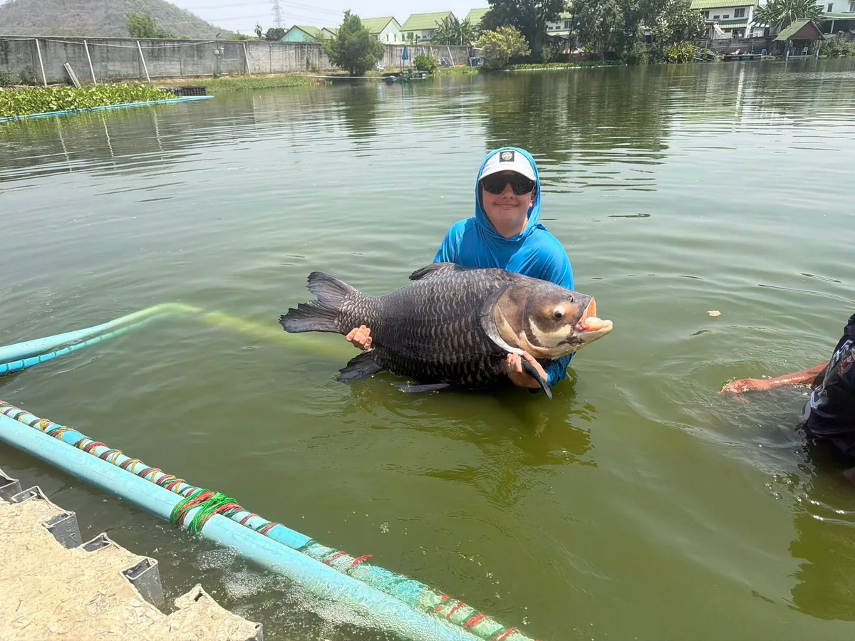 Tyler in a blue fishing top supporting a large carp in the water at Palm Tree Lagoon.
