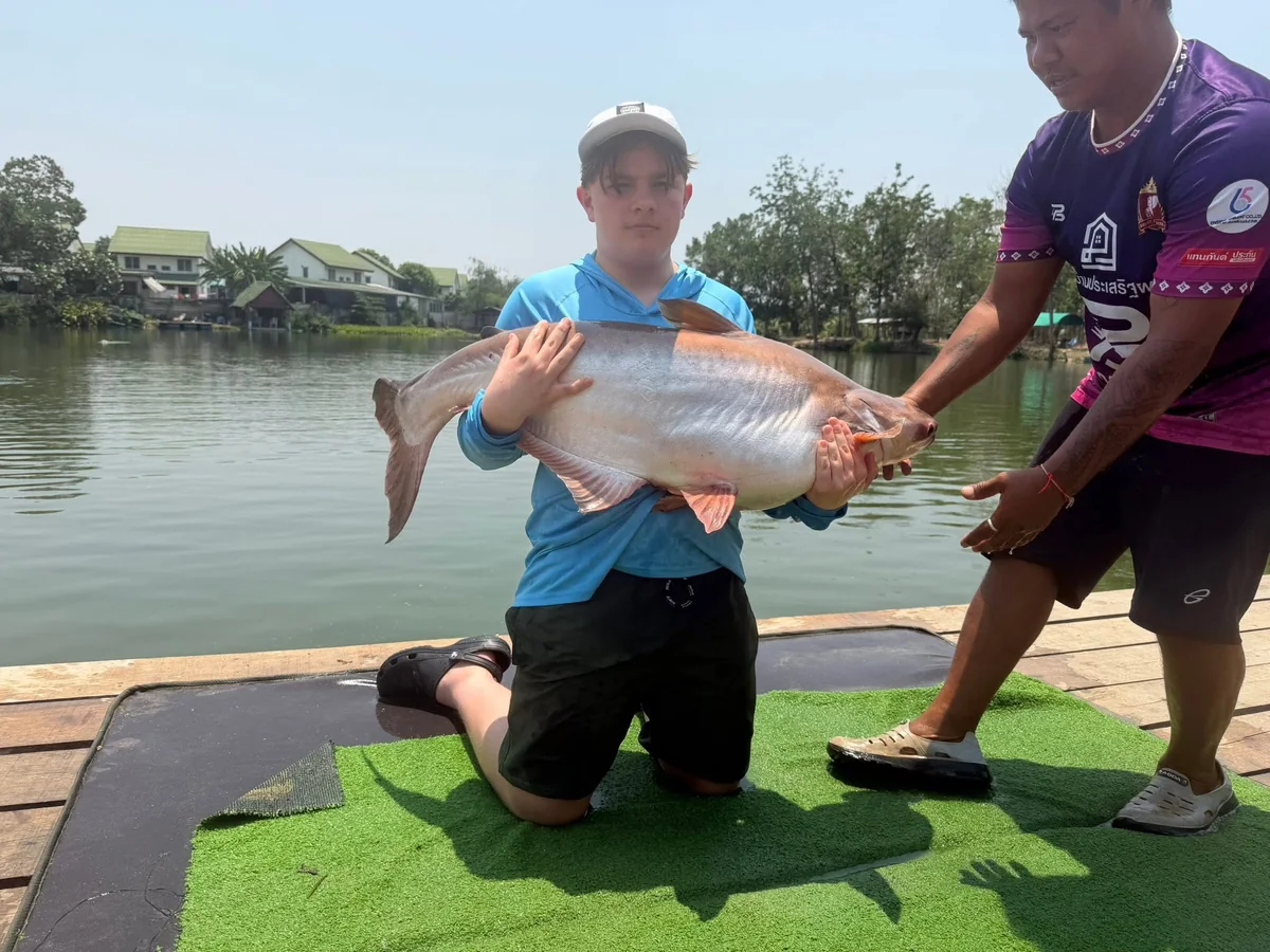 Tyler kneeling on a green mat holding a pale catfish with help from a guide.