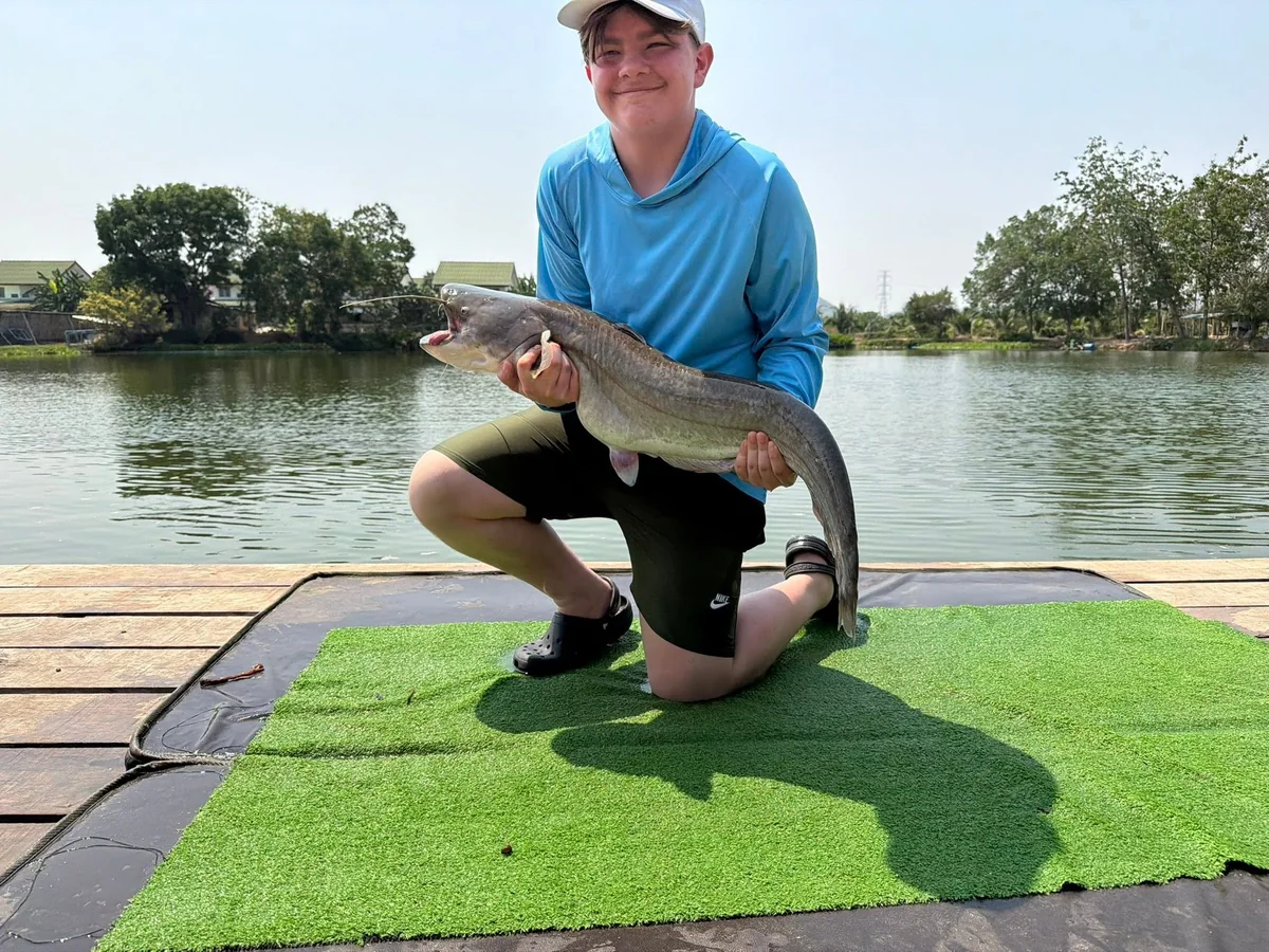 Tyler smiling and kneeling with a long grey catfish beside Palm Tree Lagoon.
