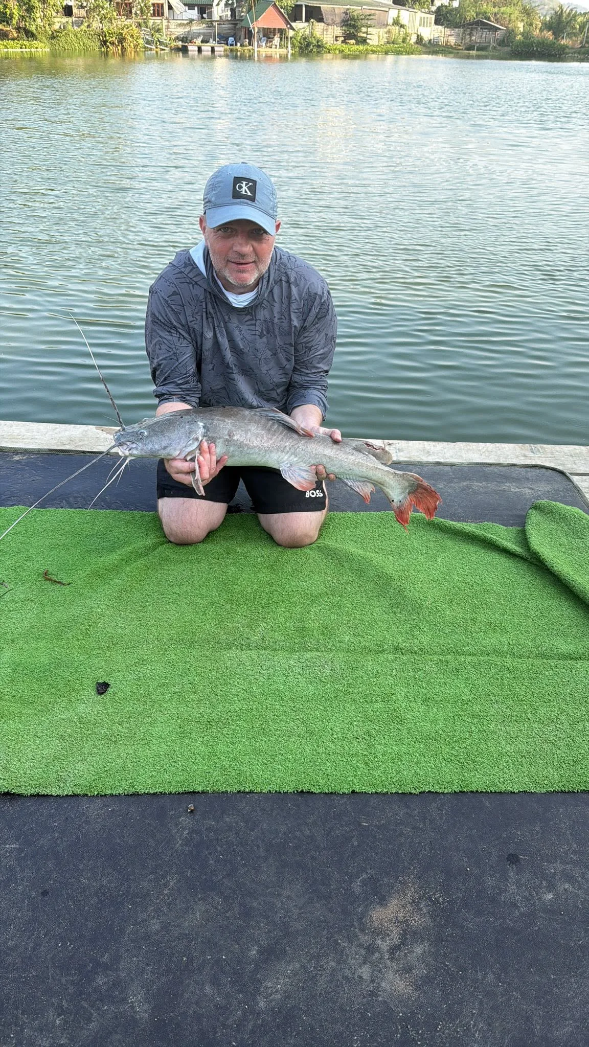 Vinny kneeling on a green mat with a red-tailed catfish beside the lake.