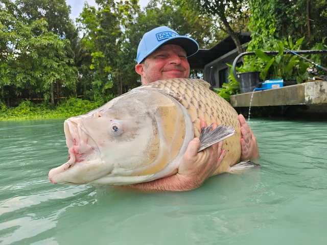 Vinny in a blue cap supporting a pale carp beside a fishing platform.